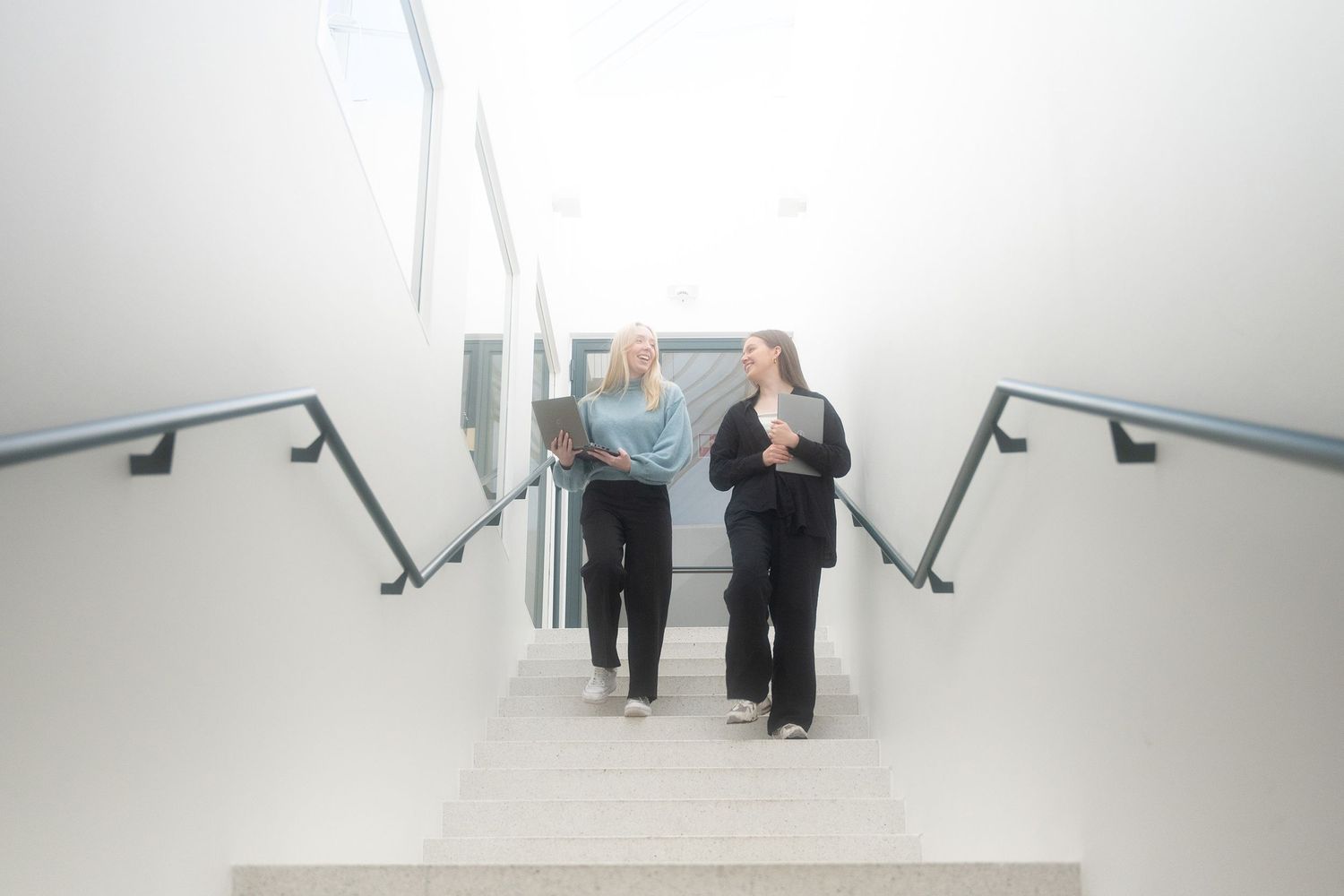 Two figures ascending a brightly lit stairwell with modern handrails.