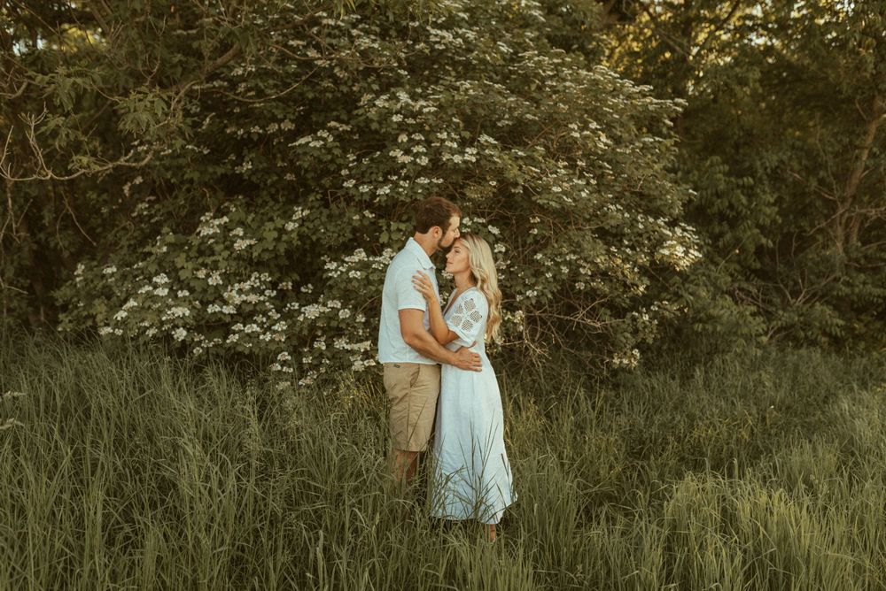 A romantic couple's portrait in tall grass with flowering trees in the background.