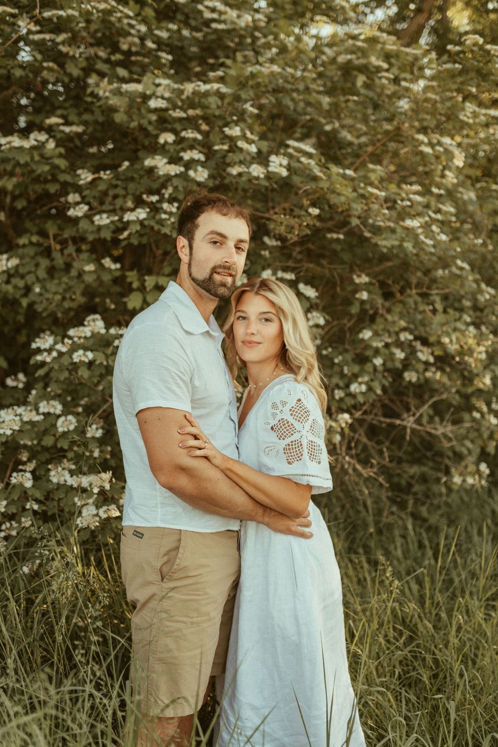 A romantic engagement photo of a couple embracing in front of flowering trees at golden hour.
