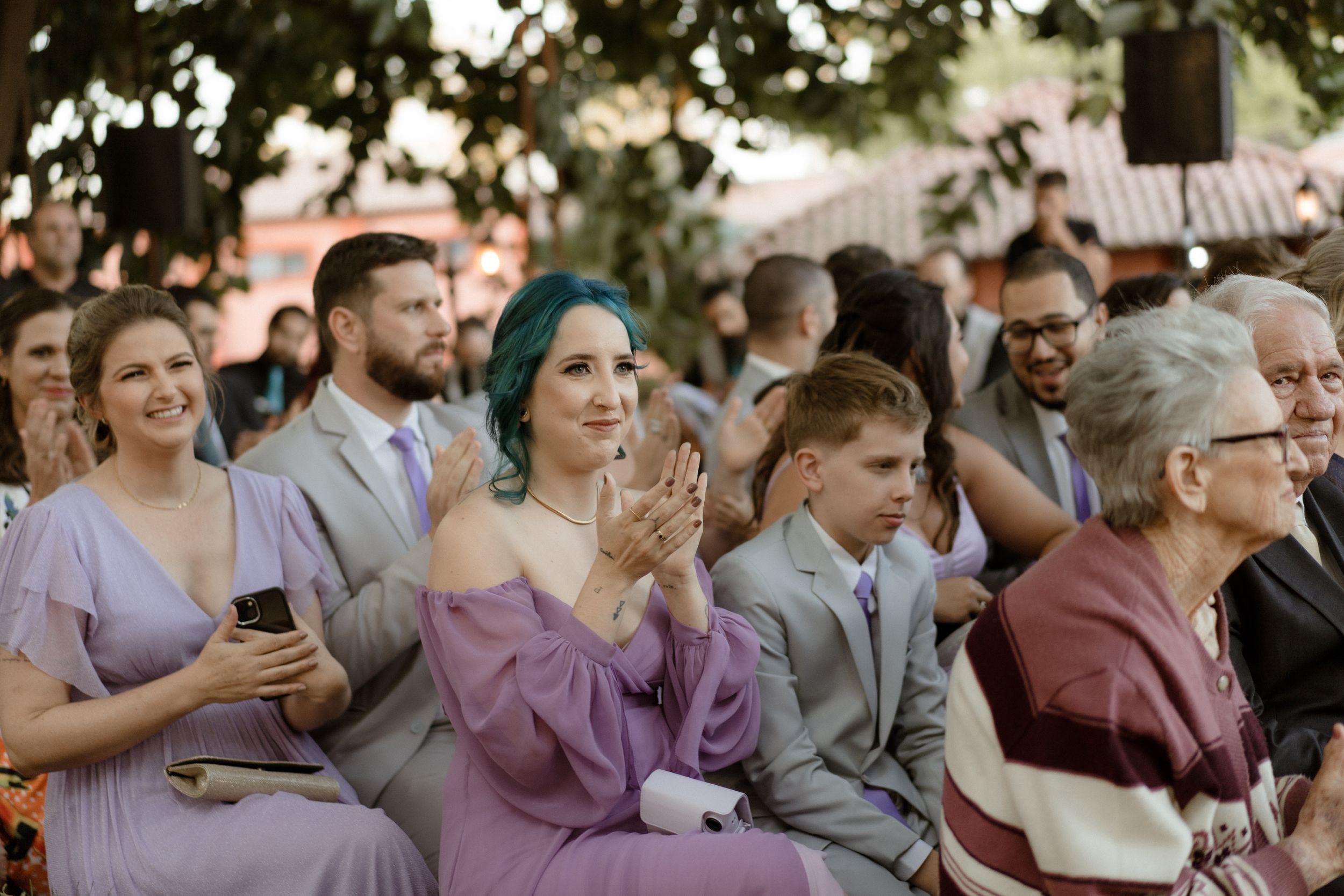 Wedding guests in purple and gray attire watch the ceremony from their seats outdoors.