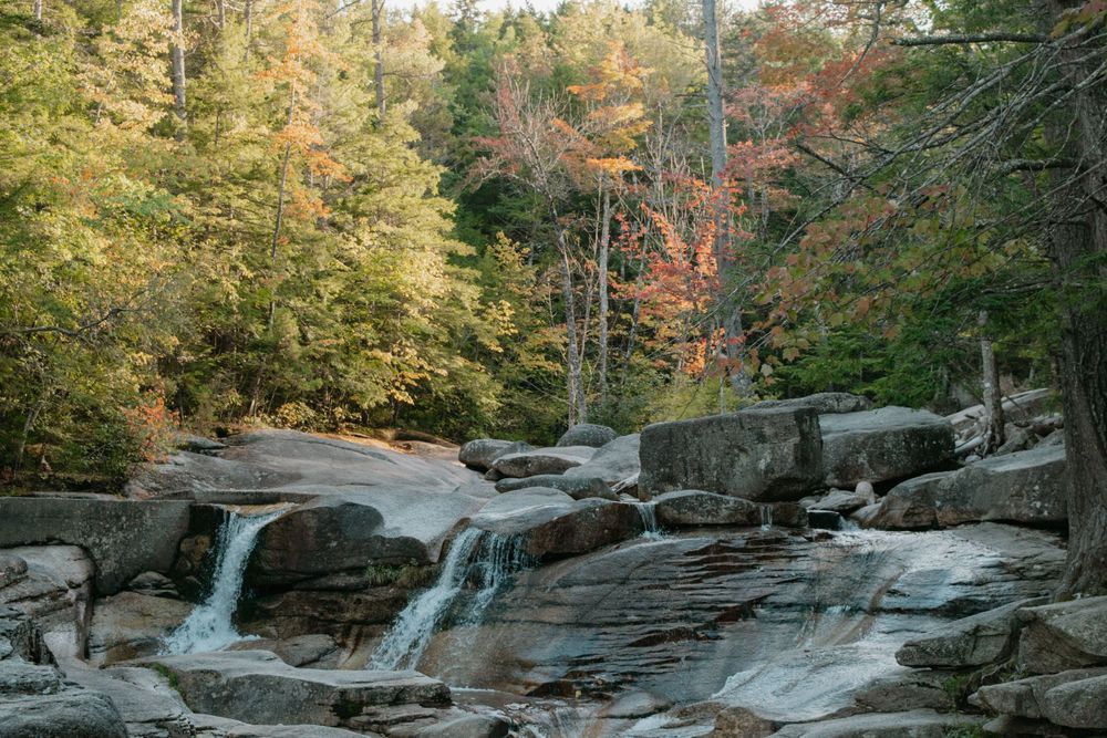 A waterfall cascades over rocky ledges surrounded by autumn foliage in a scenic forest setting. Diana's Bath's, New Hampshire