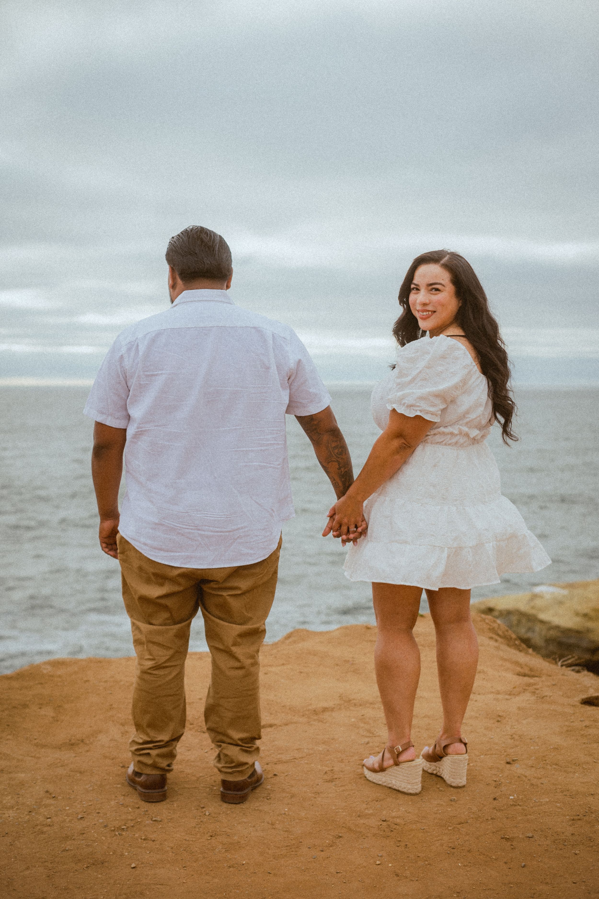 Couple in white and khaki walking hand in hand on coastal cliff during maternity shoot.
