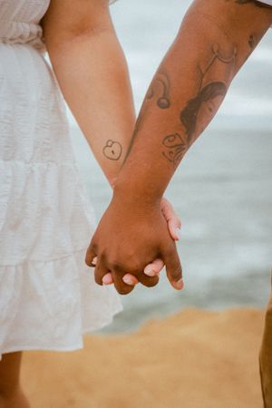 Two hands with tattoos holding each other against a sandy beach backdrop.