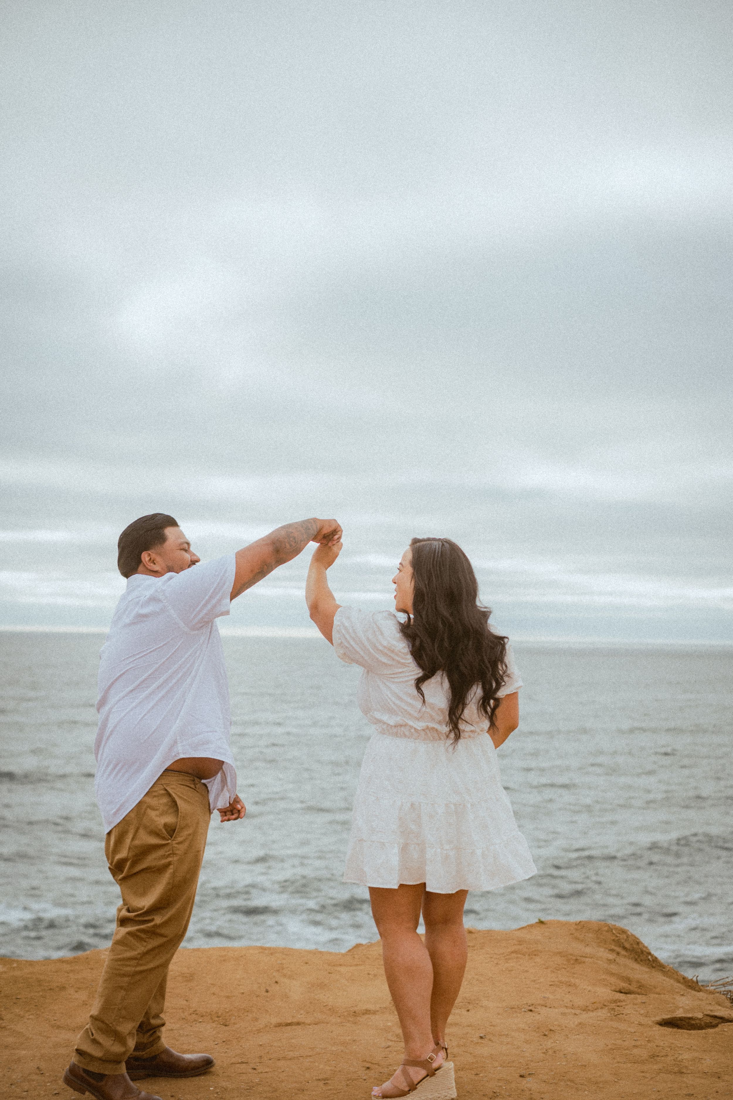 Playful couple dances together during beach maternity photoshoot against cloudy sky.