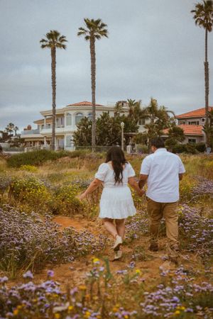 Couple walking through a field of wildflowers with palm trees and coastal homes in background.