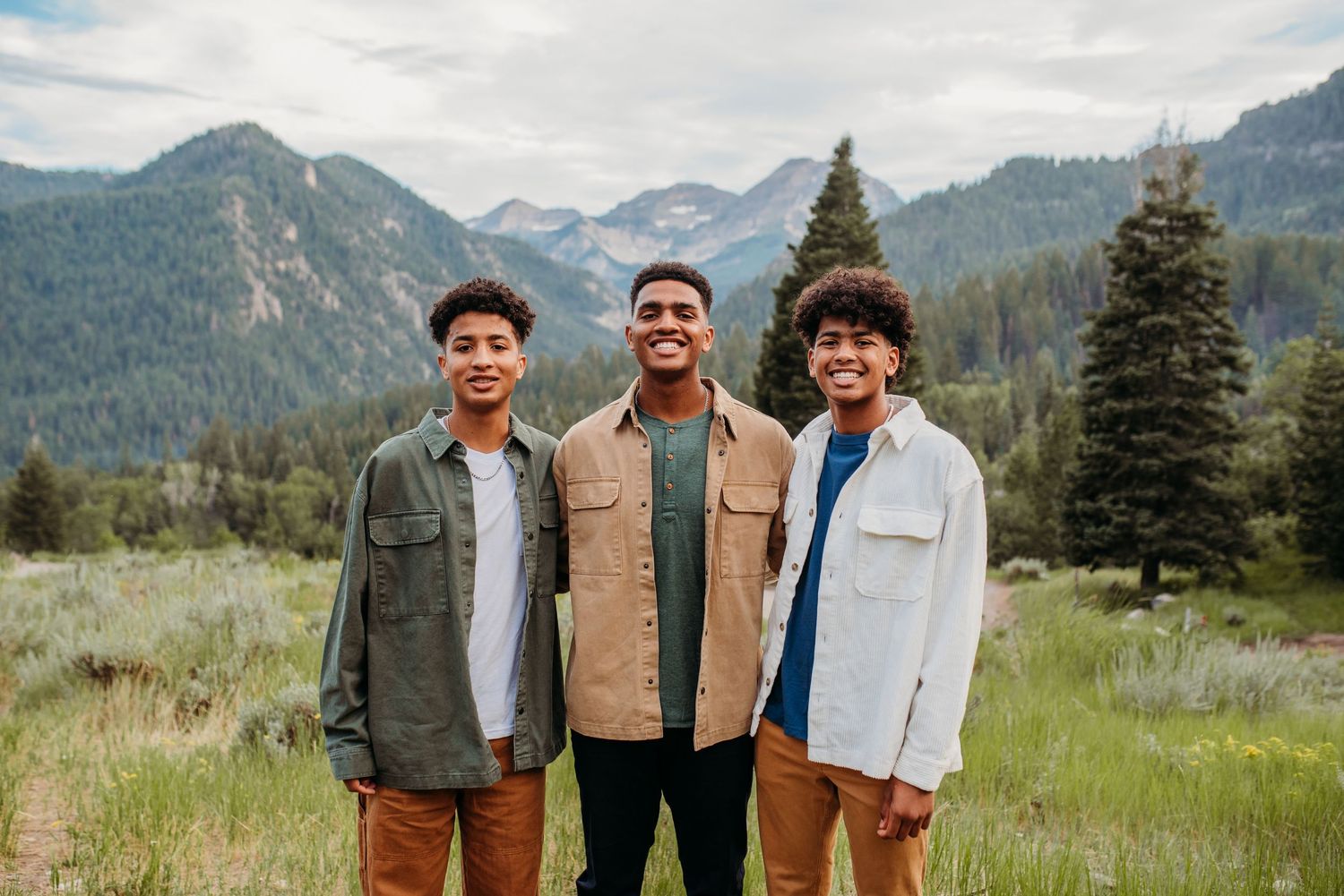 Three friends smile together with mountain peaks and evergreen forest in background.