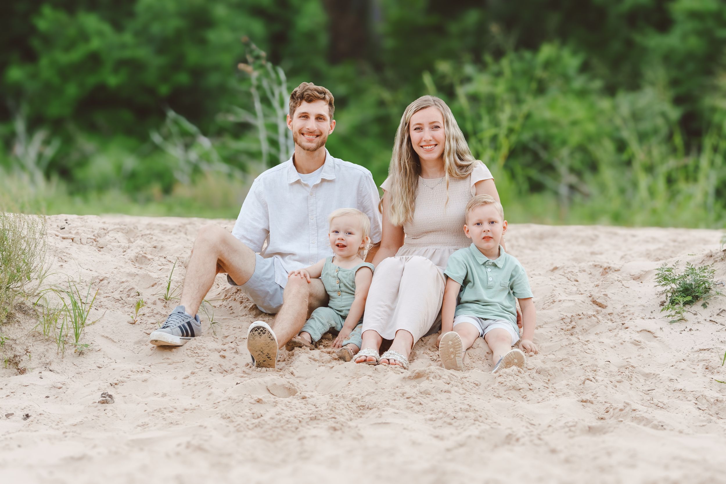 A young family sits together on a sandy beach surrounded by green foliage on a sunny summer day.