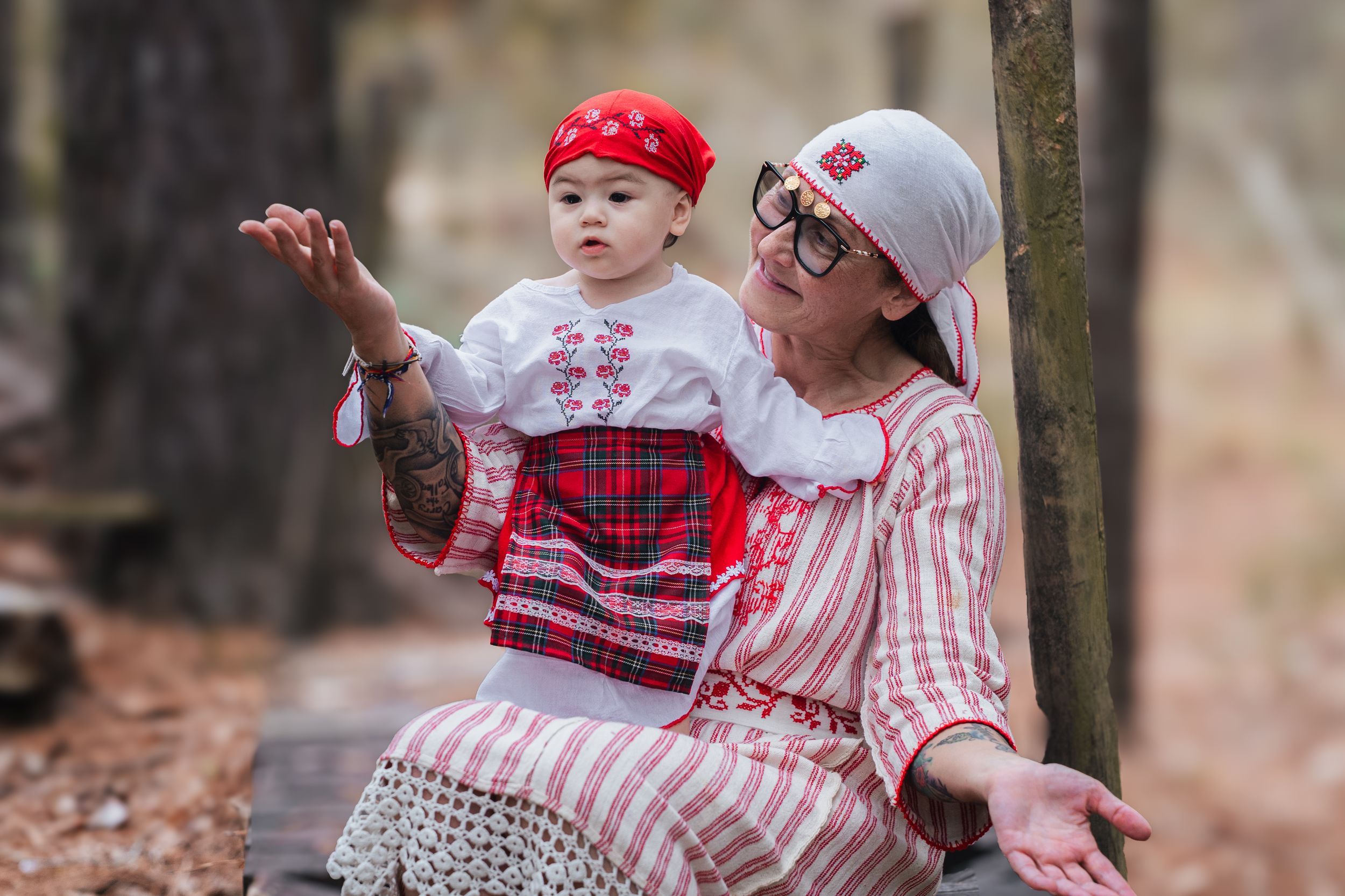 A person and baby in matching red and white outfits sit together on a log in a forest setting.
