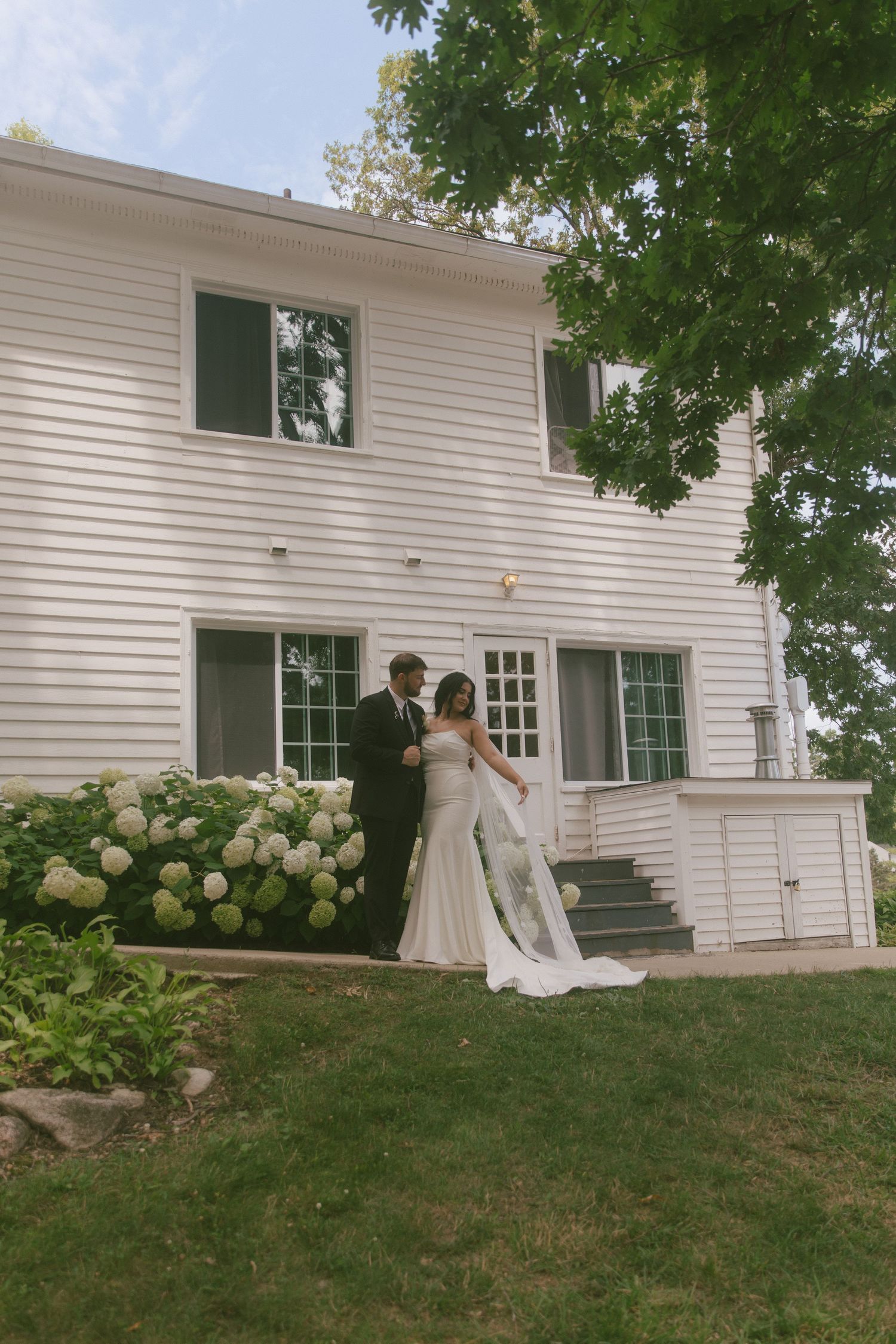 A wedding couple poses outside a white colonial house with blooming hydrangea bushes.