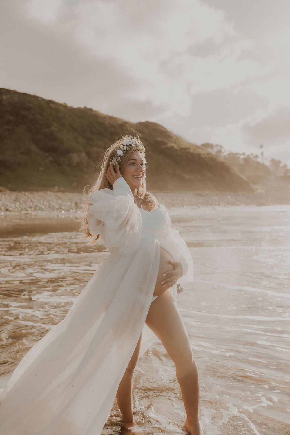 A serene beach maternity portrait with flowing white fabric against coastal cliffs.