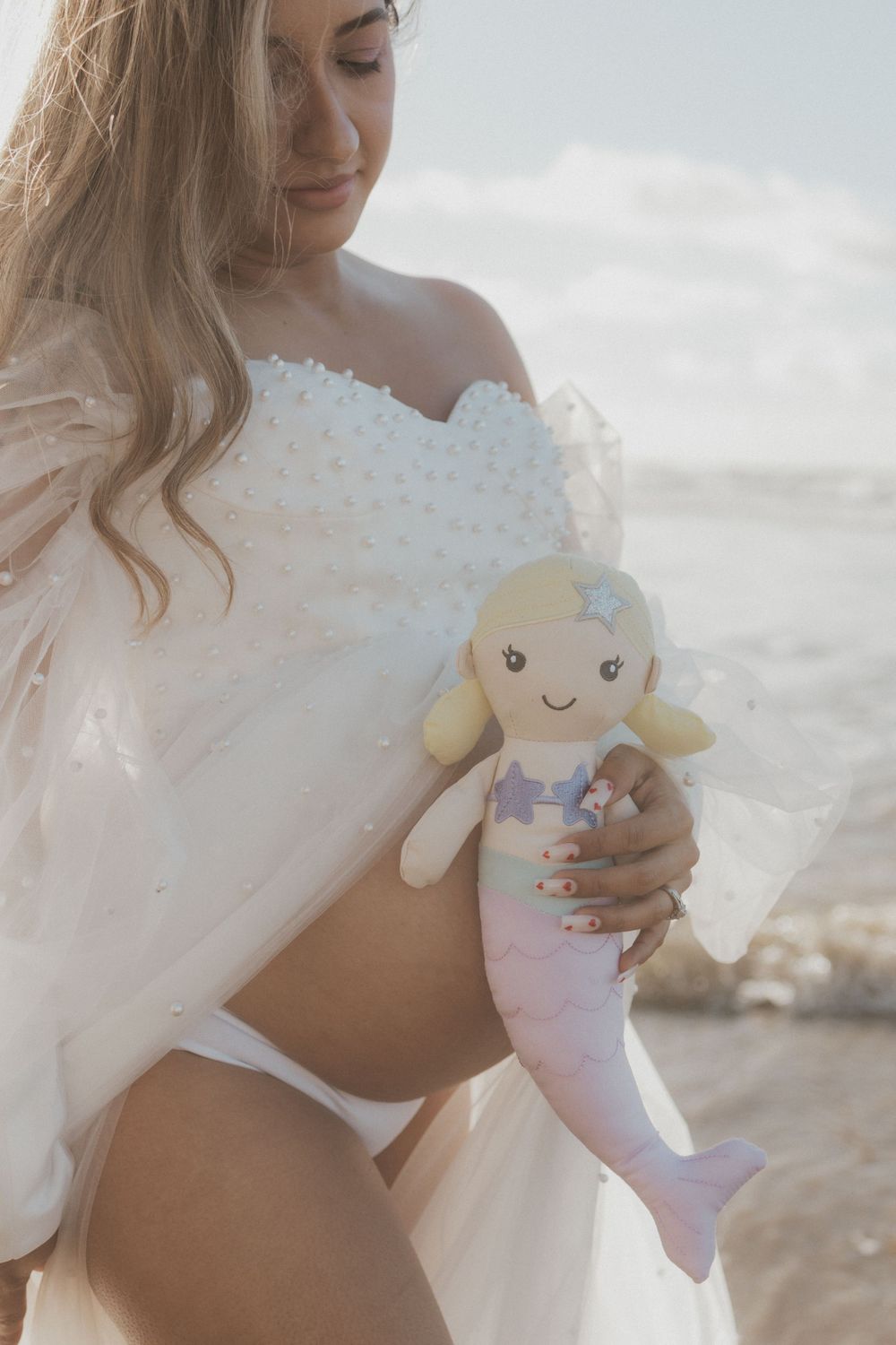 Maternity photo at beach showing white lace dress and holding cute mermaid plush toy.