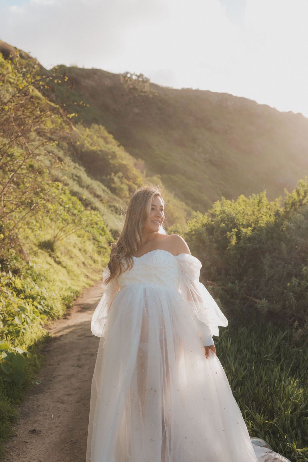 Dreamy maternity photo on mountain trail wearing flowy white gown at sunset.