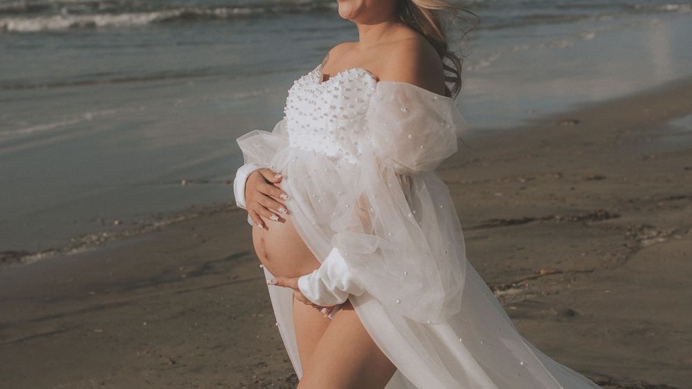 An expectant mother poses in a flowing white gown on the beach.