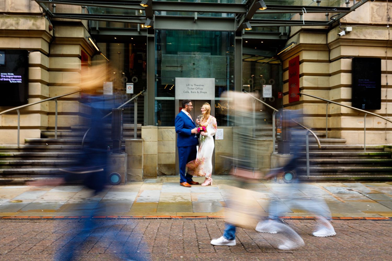 Wedding couple embraces in city doorway while blurred pedestrians walk past on brick sidewalk.