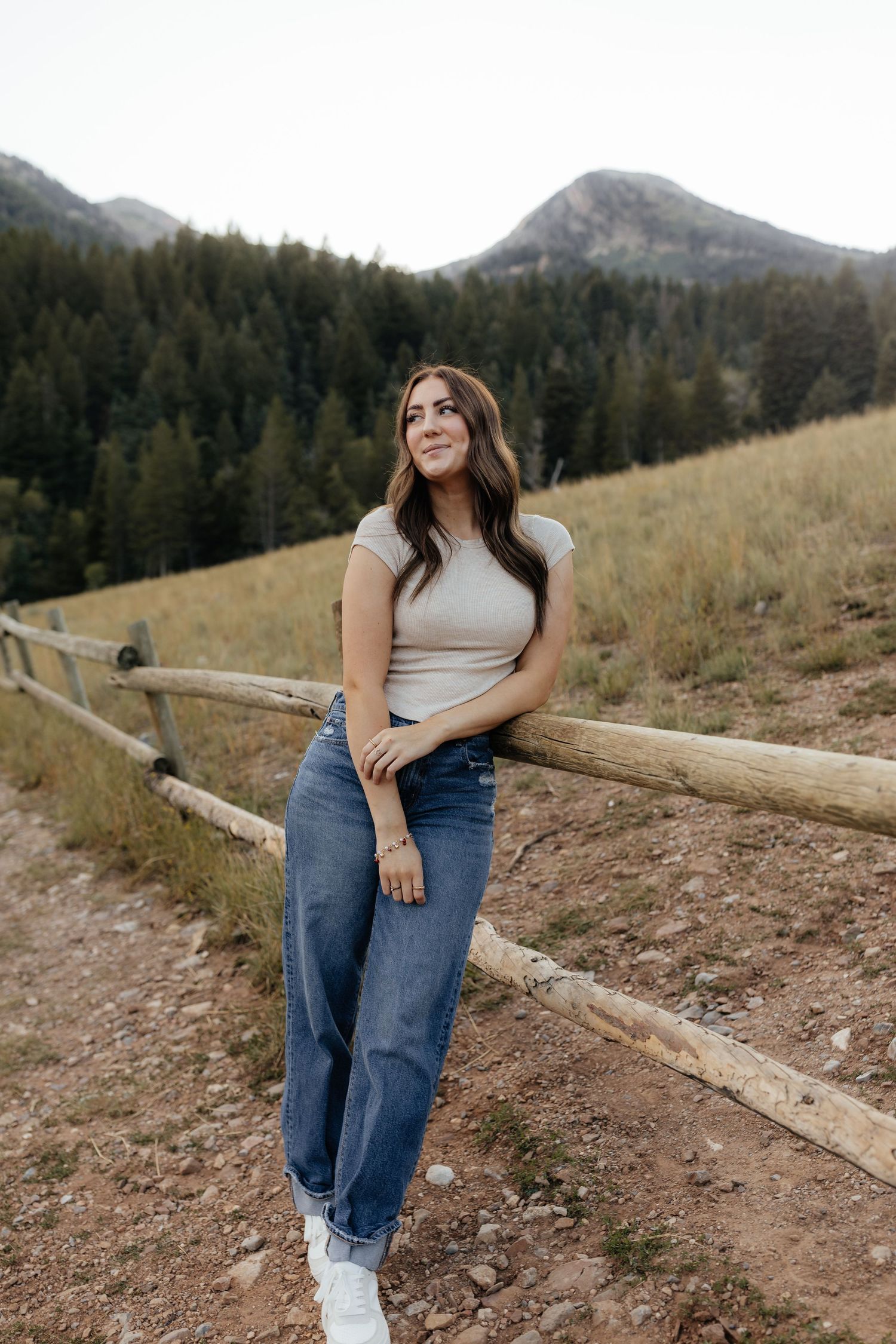 Brunette woman in a beige fitted tee and wide-leg jeans leans against a wooden fence with a forested mountain backdrop.