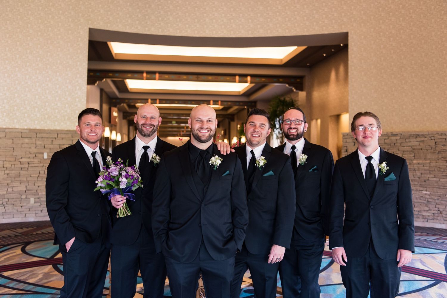 A group of groomsmen in black suits pose together at an indoor venue with boutonnieres.