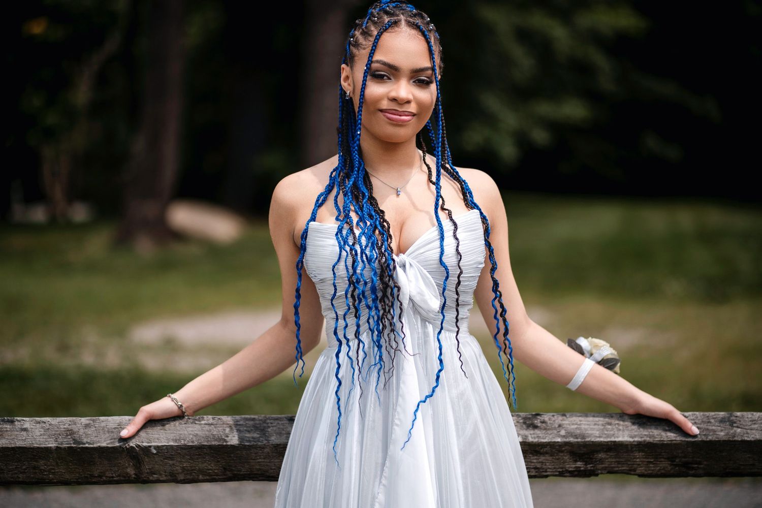 A young woman in a white dress with long blue braids poses outdoors at a scenic wooden bridge.