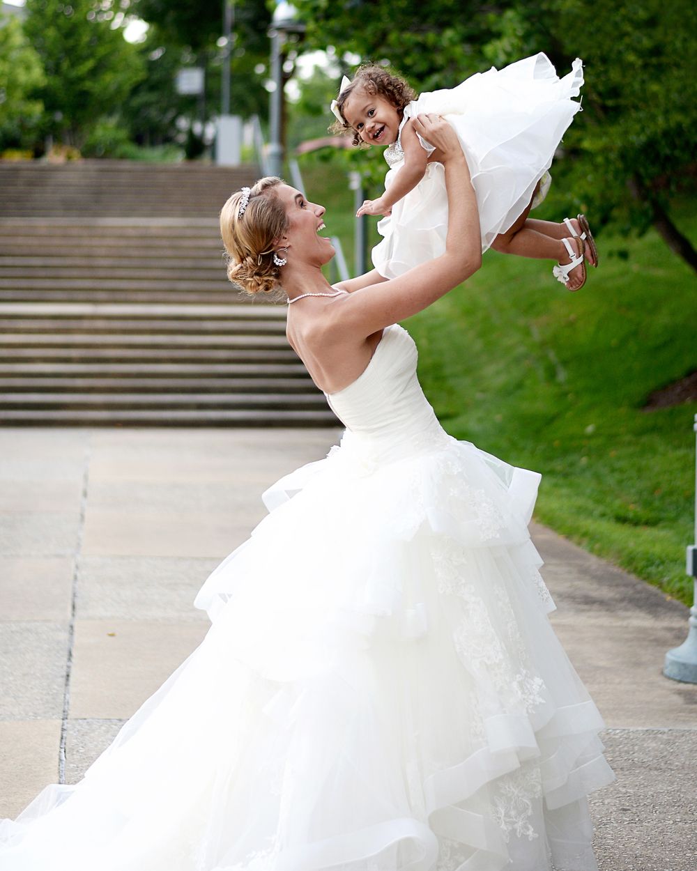 A bride in a flowing white gown lifts a small child in a white dress on outdoor steps.