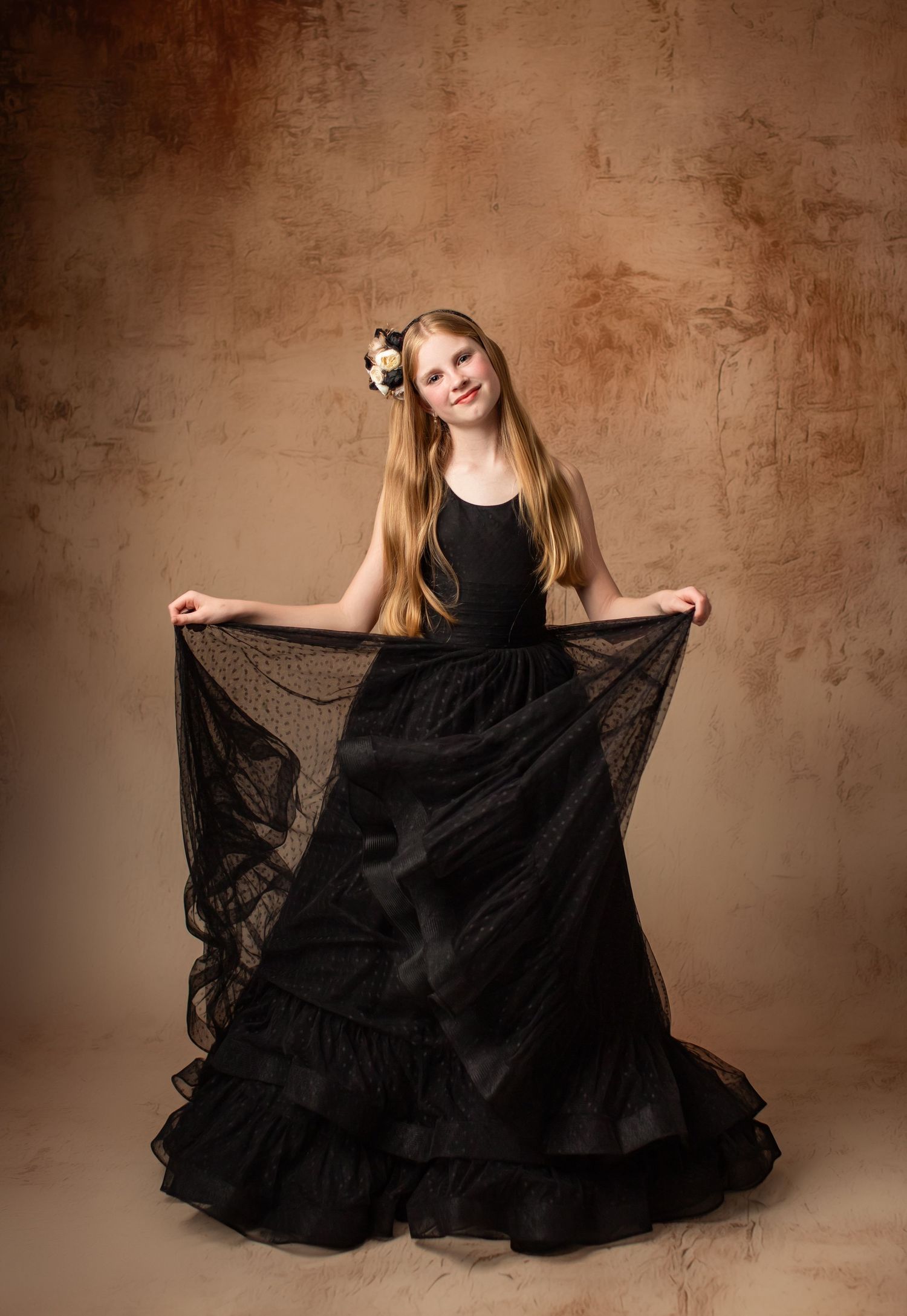 Young dancer in black flowing gown with tulle skirt poses elegantly against textured brown backdrop.