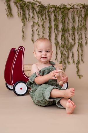 First birthday milestone session of a sweet boy in green corduroy overalls with a greenery and red wagon in foreground. Wrightstown NJ