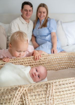 A shot of a big sis looking into a Moses basket of her new baby sibling swaddled. Parents in foreground smiling on at the sweet moment.
