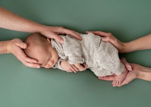 A close up shot of a newborn with parent's hands on their hand and legs/bum. Parent at each end of baby with just hands in shot.
