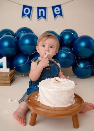 Cake smash session with a sweet 1 year old in denim overalls enjoying his cake with balloons and a ONE banner in the background.