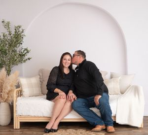 A couple in studio with a white arch backdrop sitting on a daybed as the husband leans over and kisses wives cheek. Wrightstown NJ