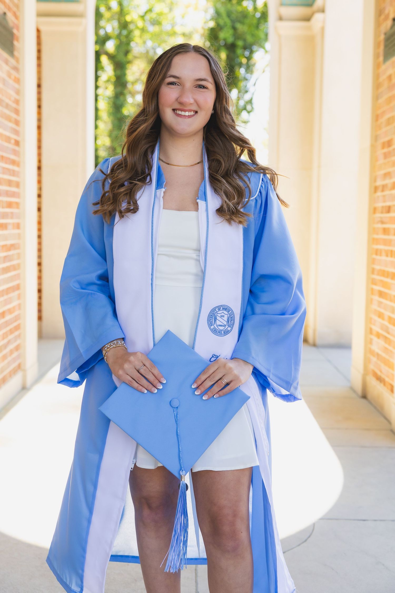 Smiling graduate in light blue cap and gown holding mortarboard outdoors.