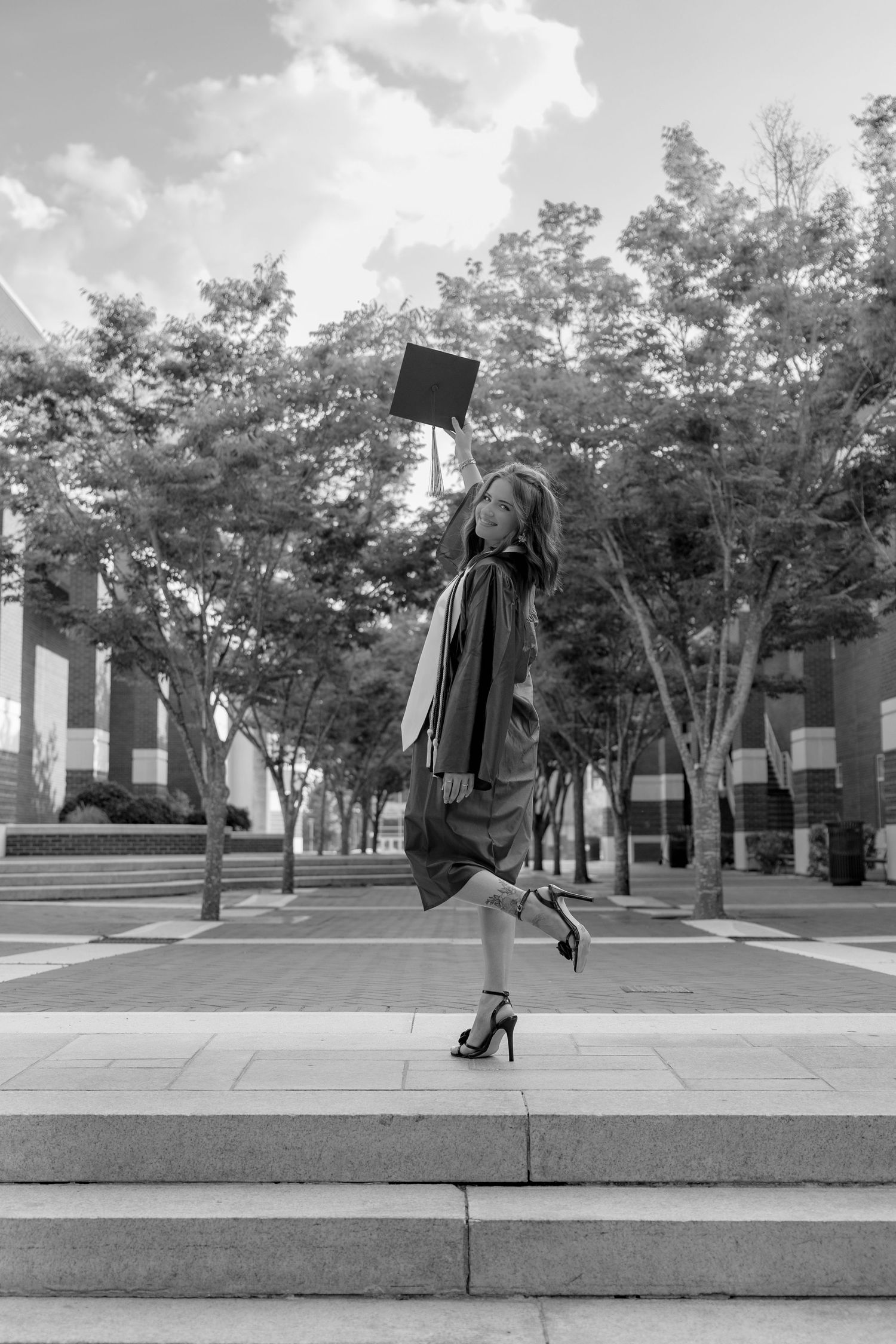 Black and white photo of a graduate in cap and gown tossing mortarboard on steps outdoors.