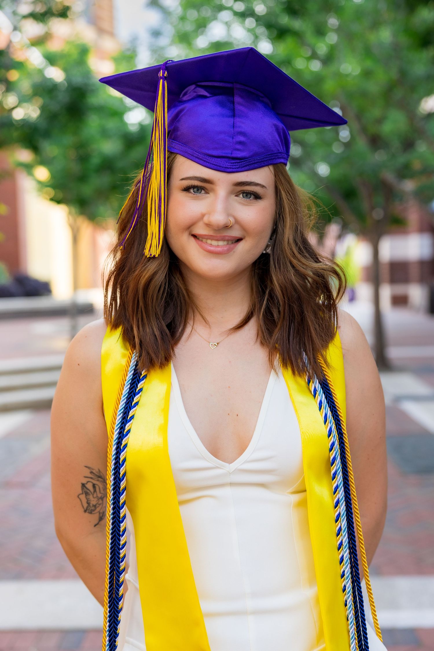 Smiling graduate in purple cap and yellow sash poses outdoors in white dress on a sunny day.
