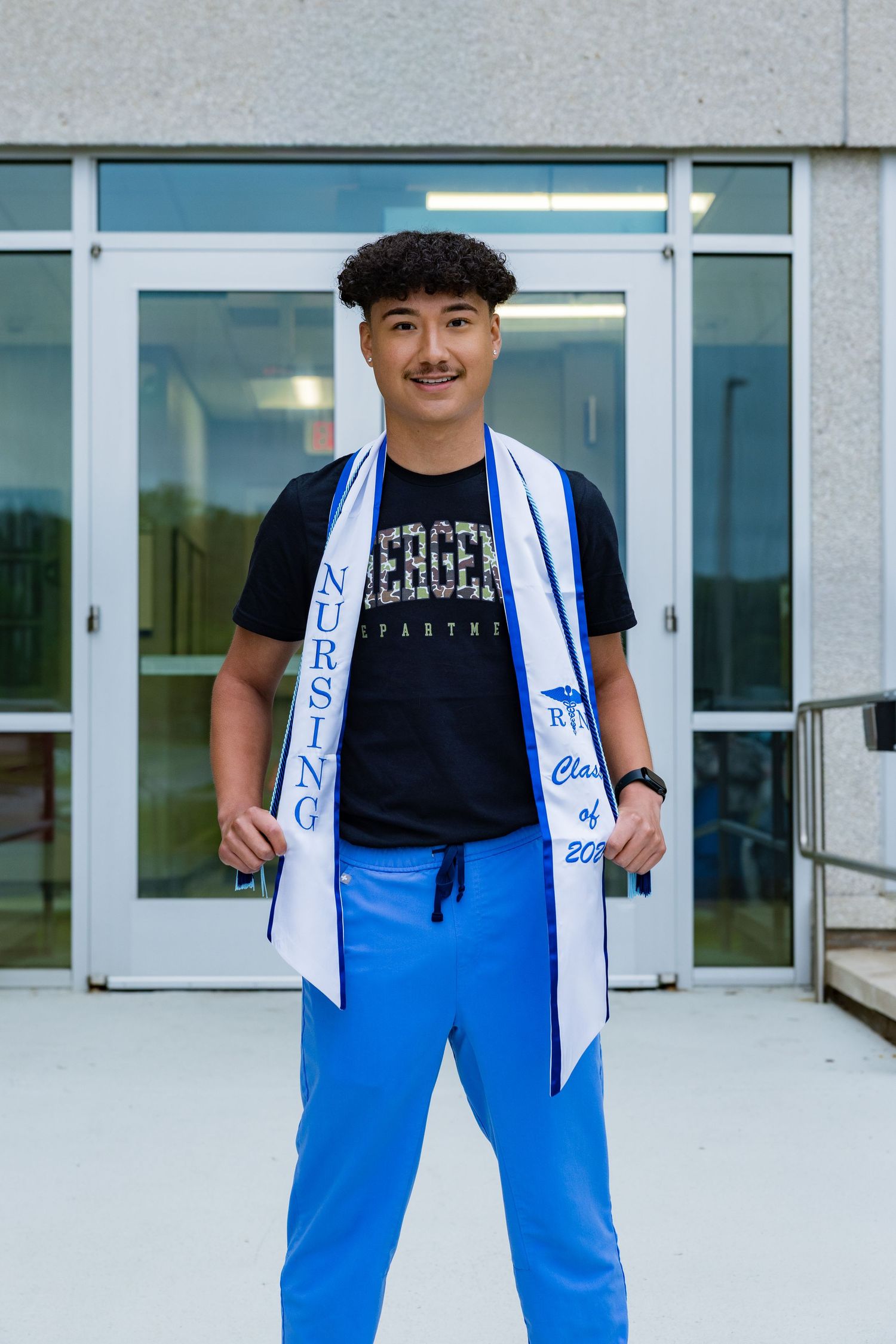 Young nursing graduate smiles proudly wearing blue scrubs and a white graduation sash outside a building.