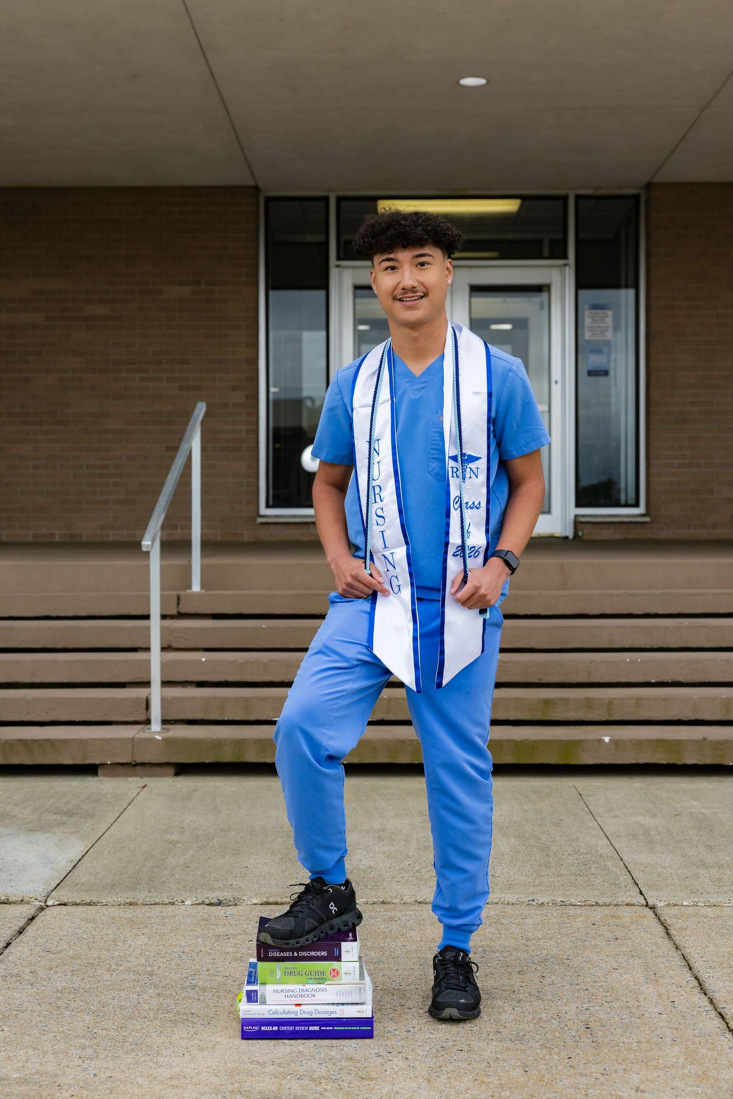 Young man in blue scrubs and graduation stole poses with medical textbooks outside a university building.