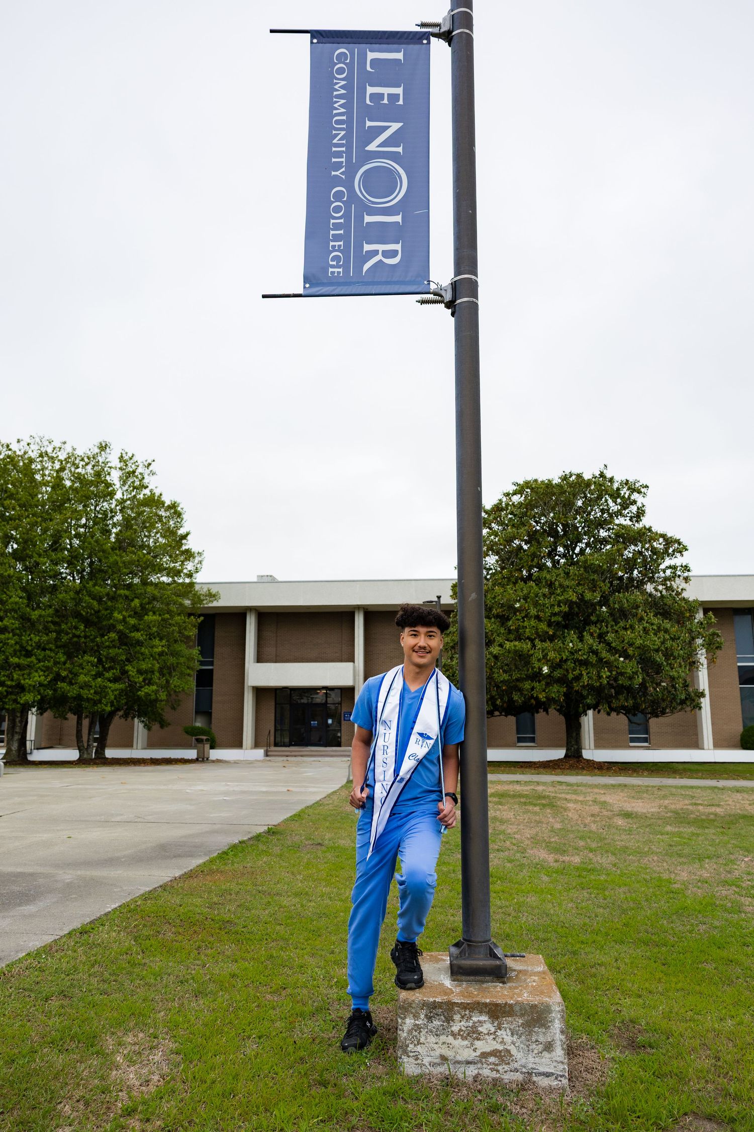 Student in blue scrubs poses by Lenoir Community College banner sign on campus grounds.