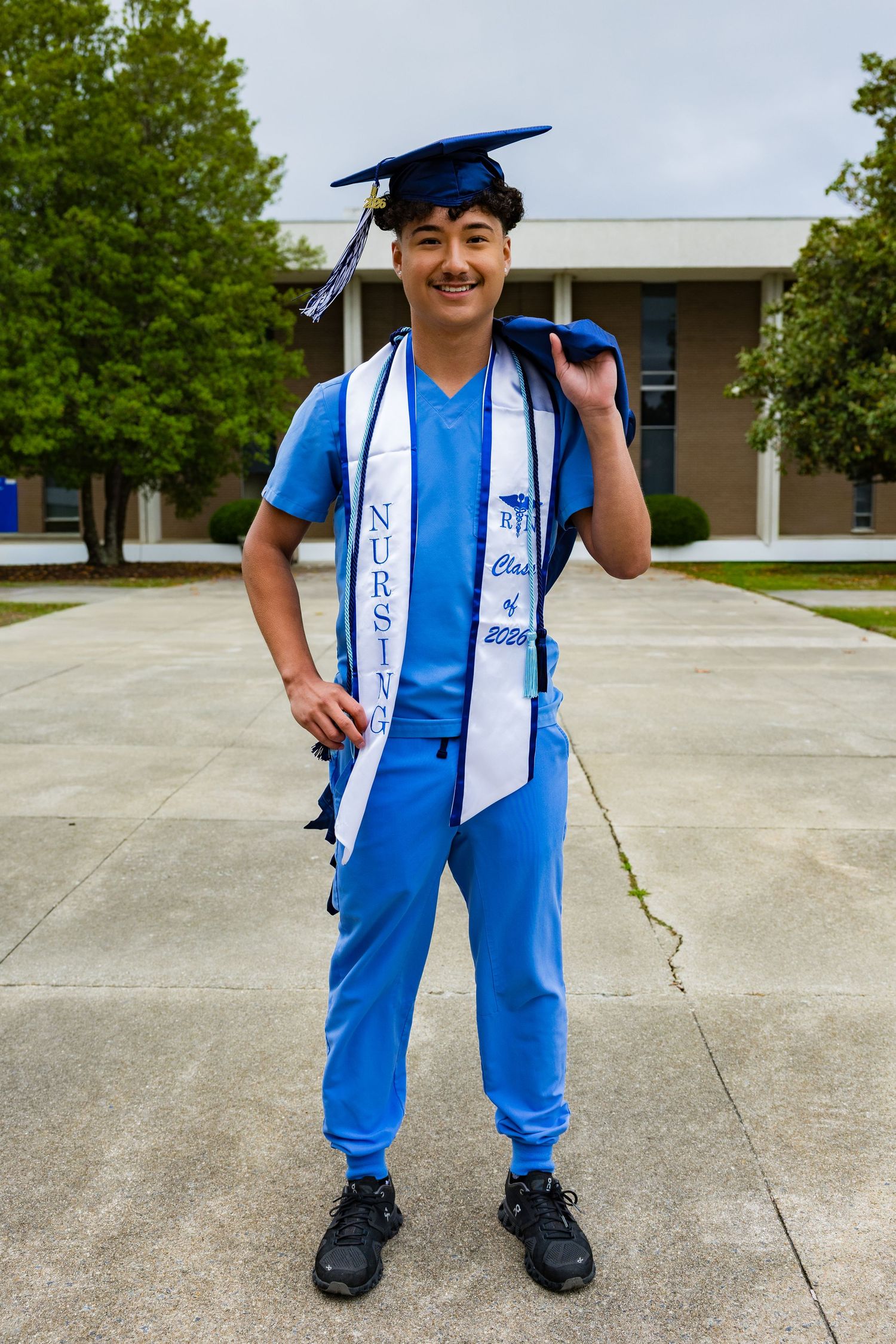 Smiling nursing graduate in blue scrubs and cap with graduation stole poses outdoors on campus.