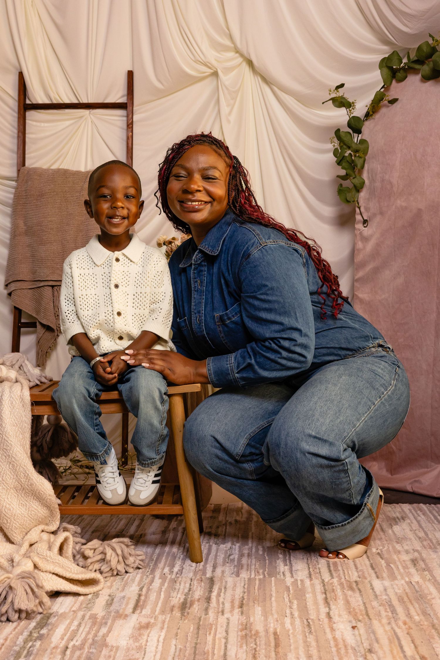 A smiling woman in denim outfit crouches beside a young boy in white shirt and jeans during a studio portrait session.