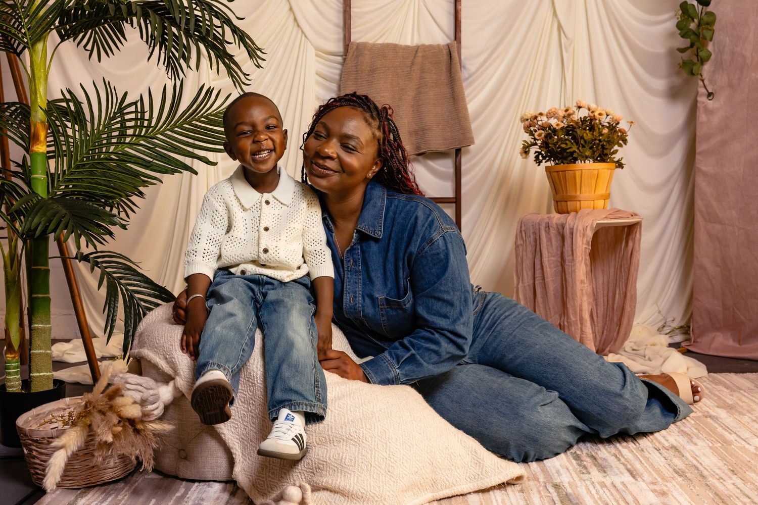 A woman and young boy smile together on floor cushions surrounded by plants in a cozy studio setting.