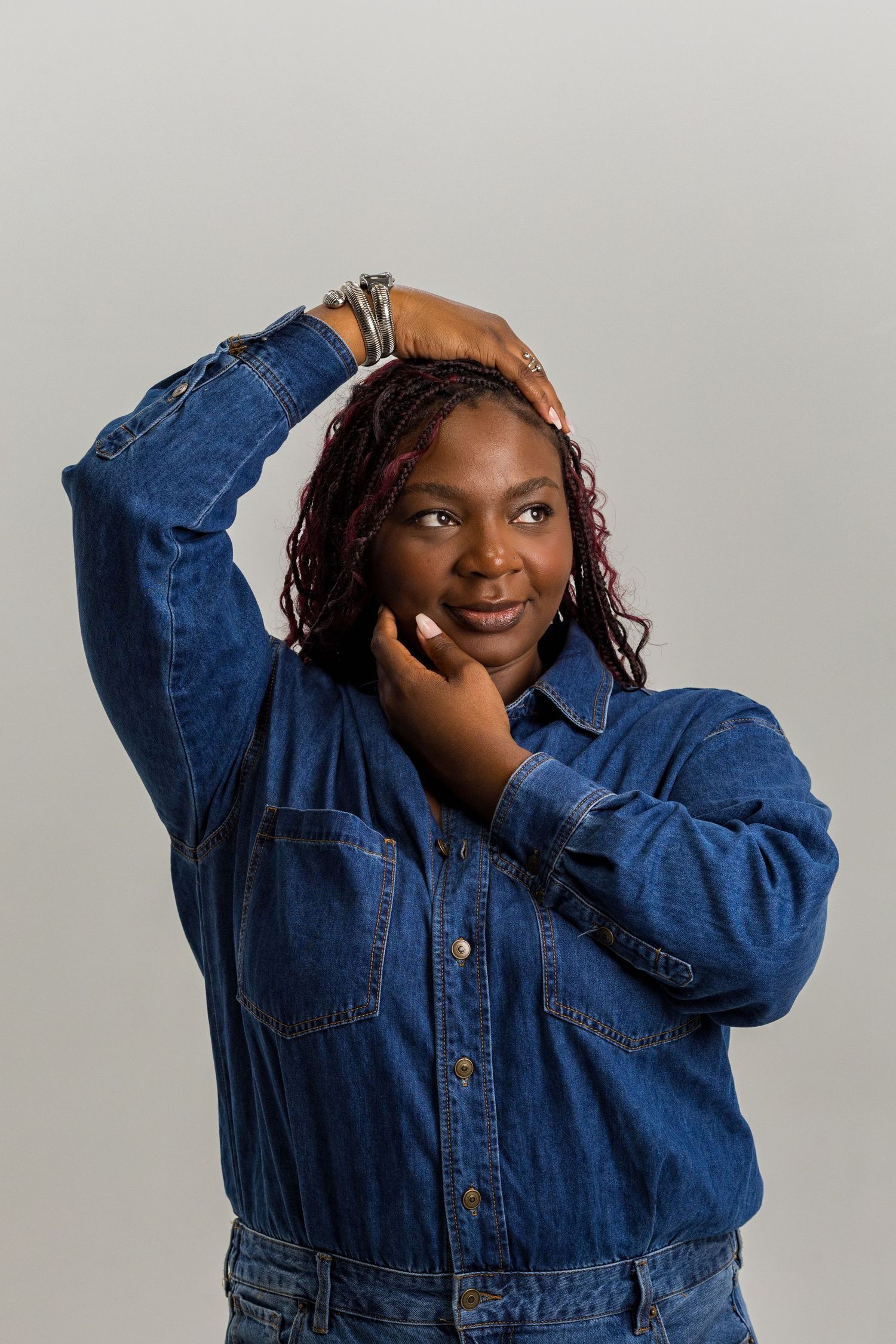 A woman in a denim jacket poses with hands near her face and head, wearing braided hair and silver bracelets.