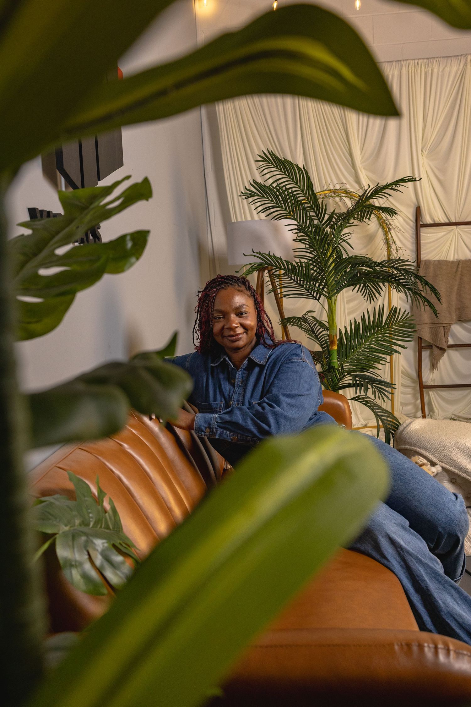 A smiling Black woman in a denim outfit relaxes on a brown leather sofa surrounded by lush green tropical plants indoors.