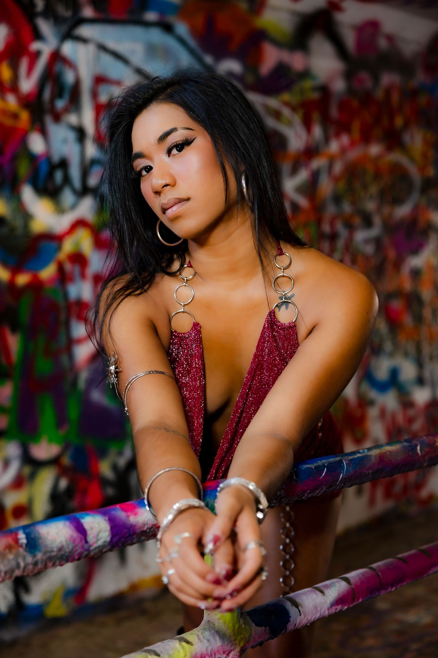 Young woman in burgundy top poses against colorful graffiti wall, wearing hoop earrings and silver bracelets.