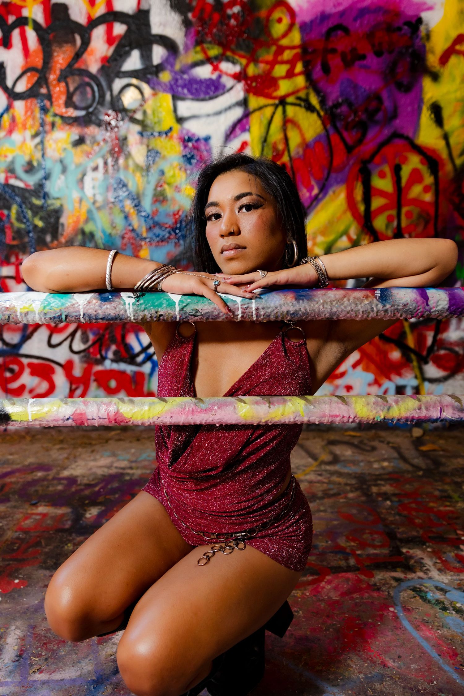 Young woman in red dress poses kneeling against a colorful graffiti wall, resting arms on a painted bar.