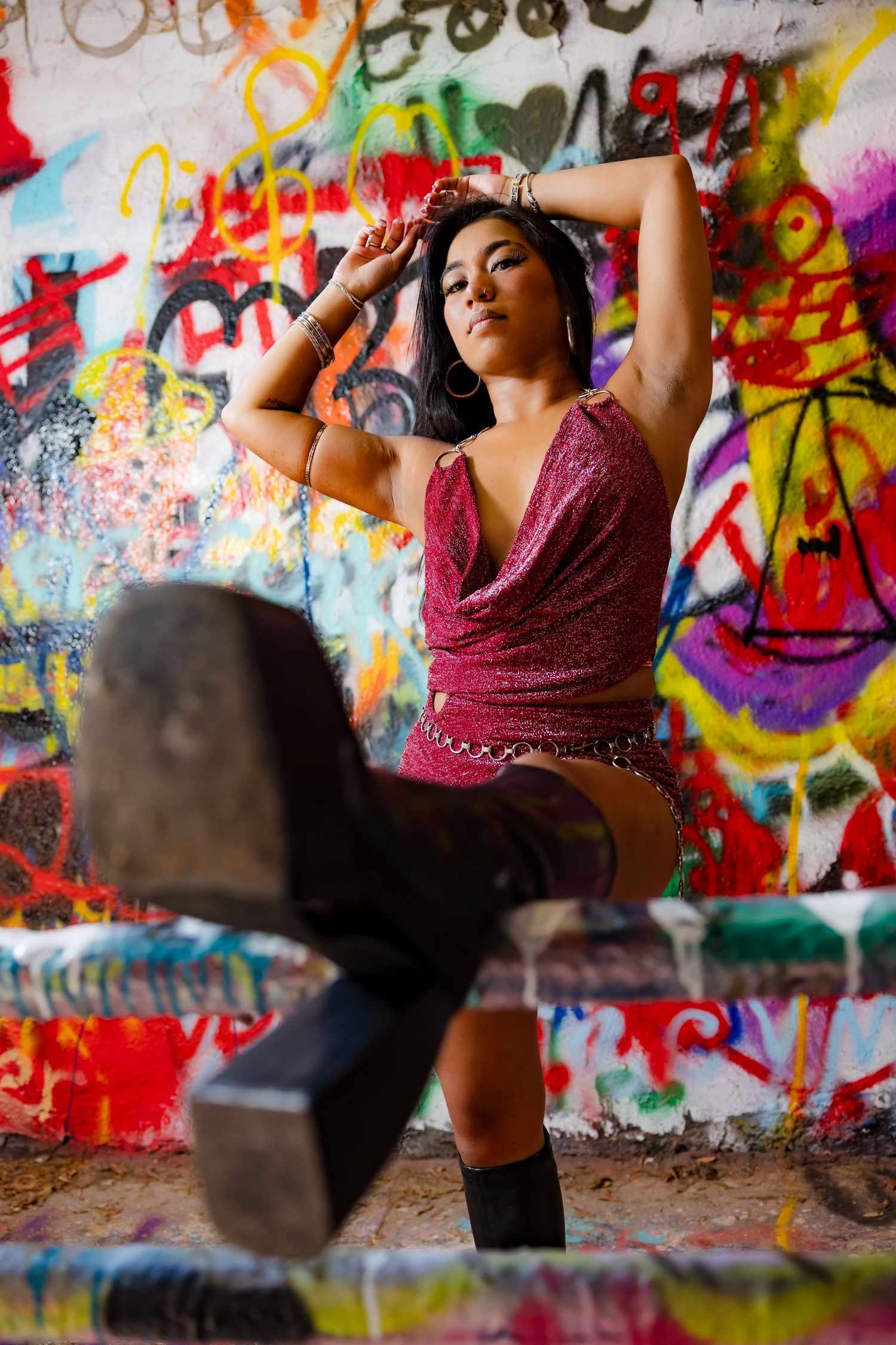 A woman in a red sparkly outfit poses confidently in front of a colorful graffiti wall, with a boot raised in the foreground.