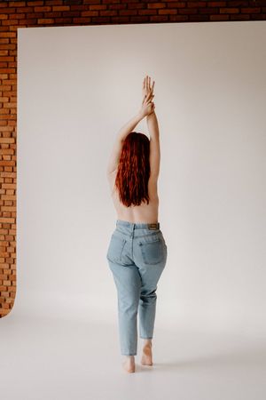 Redhead woman posing topless in blue jeans with arms raised, shot from behind against white studio background.