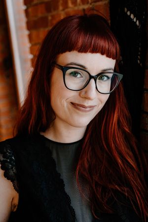 Woman with red hair, bangs, and black cat-eye glasses smiling warmly in front of a brick wall.