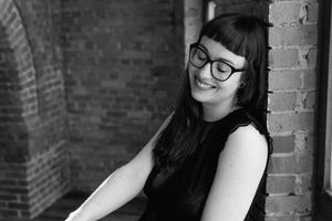 Black and white portrait of a smiling woman with glasses and dark hair leaning against a brick wall.