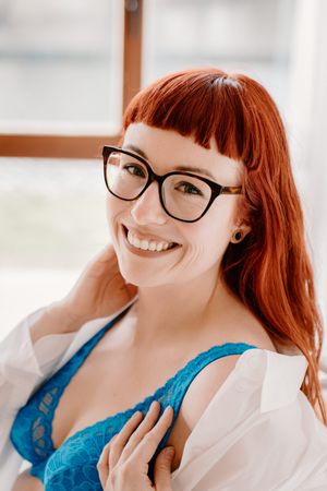 Smiling redhead woman wearing glasses and blue lace bra with open white shirt in a bright indoor setting.
