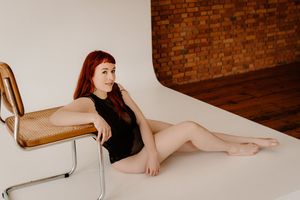 Young redhead woman in black bodysuit posing on floor beside vintage chair in photography studio.