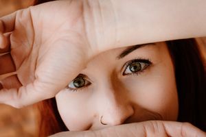Close-up of a young woman with green eyes and a nose ring, framing her face with her hands.