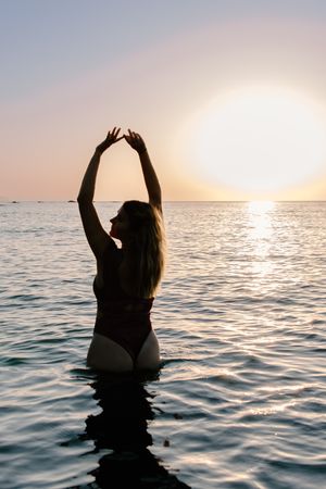 A silhouetted woman stands in the water with her arms raised at sunset, creating a serene and peaceful scene.