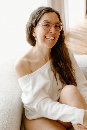 Smiling woman with glasses and long hair wearing an off-shoulder white sweater, sitting relaxed on a light sofa.
